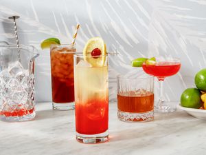 Four different cocktails with different garnishes and in different cocktail glasses with a pitcher full of ice with a bar spoon inside of it. On one side there is a bottle of liquor getting cut off and the on the other side there is a plate full of lemons and limes