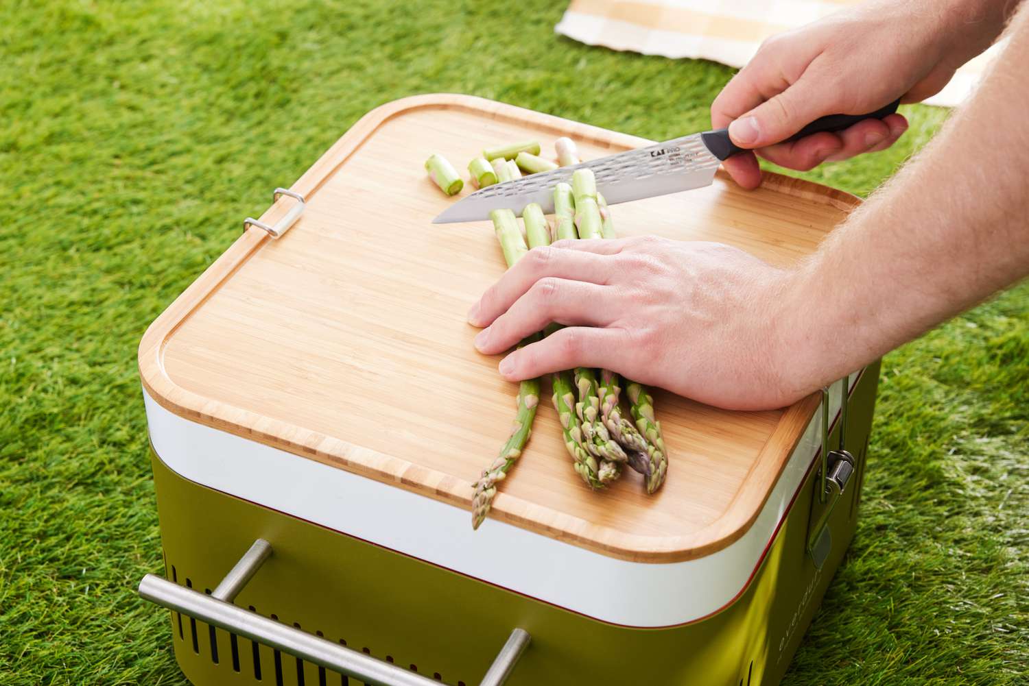 Hands chopping asparagus on a wooden cutting board on top of a Everdure Cube Portable Charcoal Grill