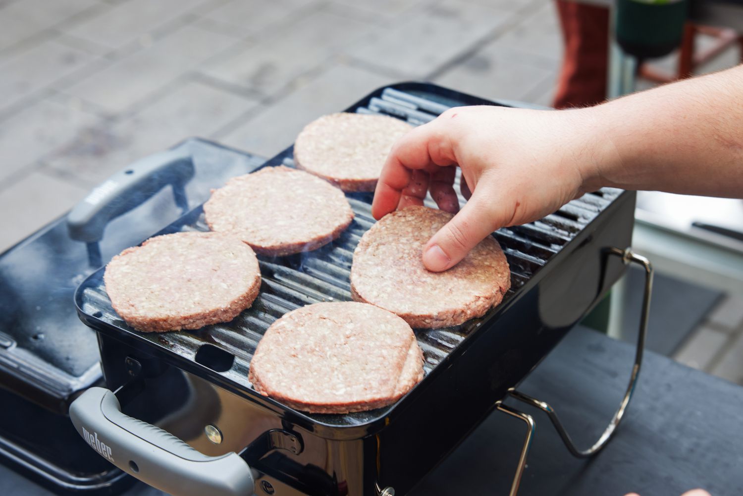 Hand placing raw burger patties on a Weber Go-Anywhere Charcoal Grill on a table 