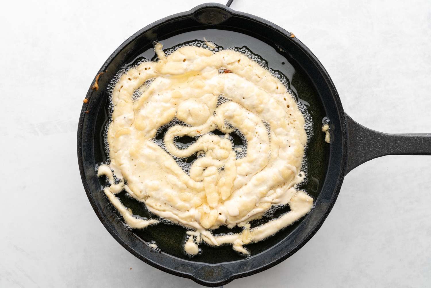 Funnel cake in a round, squiggly pattern almost entirely filling the skillet with the hot oil 