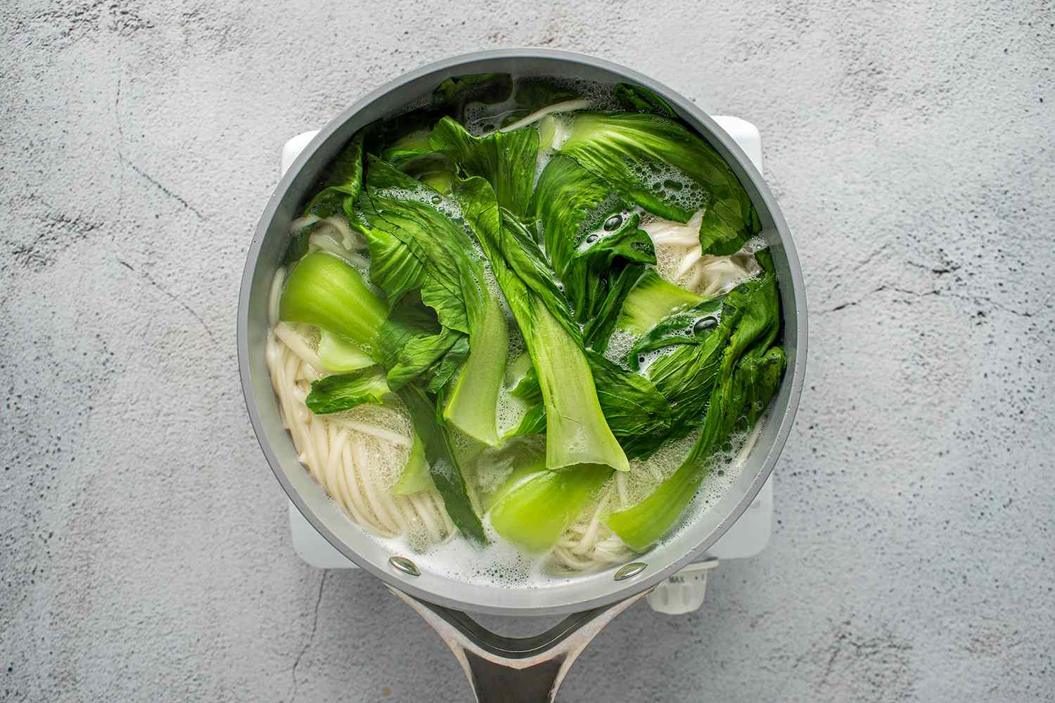 Noodles and bok choy being cooked in pot on a burner 