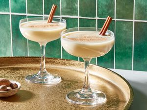 Two eggnog martinis, garnished with nutmeg and a whole cinnamon stick, on a gold tray with a small bowl of whole nutmeg