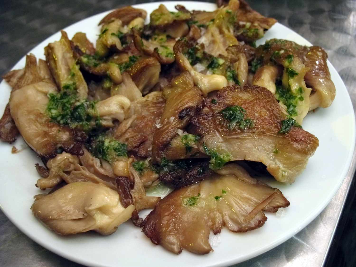 Tapas of pan-fried oyster mushrooms with salsa verde on a white plate on a stainless steel tabletop