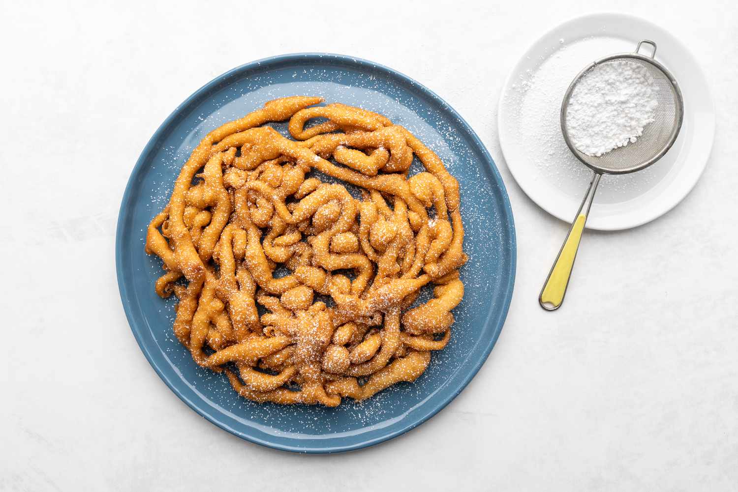 Funnel cake on a blue serving plate being dusted with confectioners' sugar with a small sieve