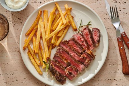 A plate of sliced ribeye steak and french fries, with a small bowl of aioli served on the side