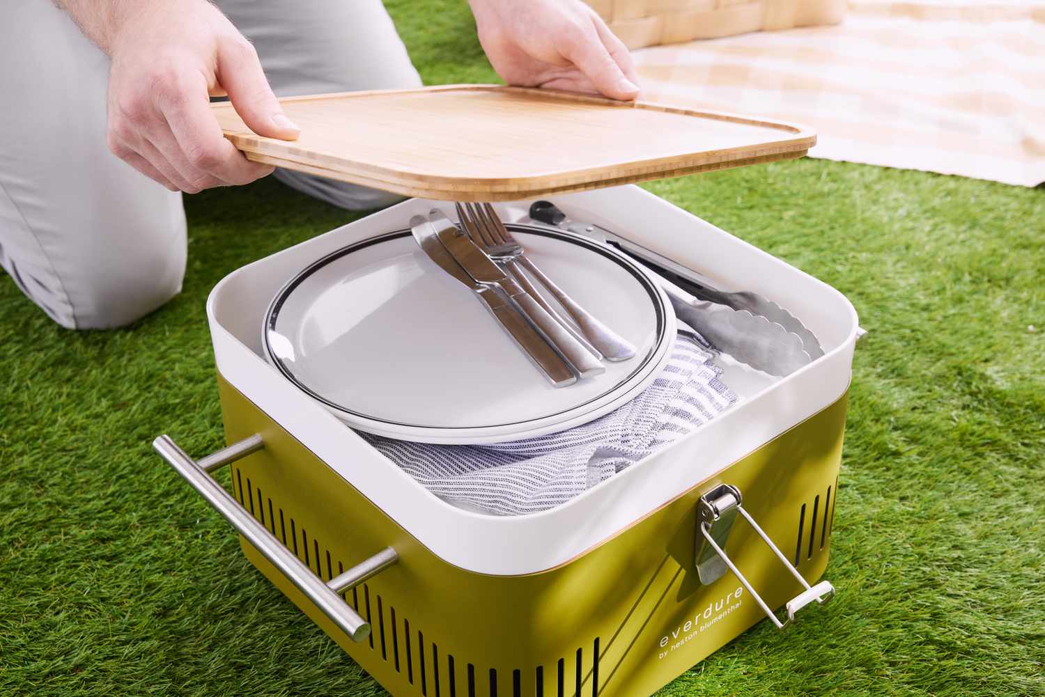 Person placing wooden cutting board on top of Everdure Cube Portable Charcoal Grill with plates and cutlery inside 