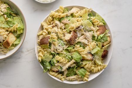 A large bowl of chicken caesar pasta salad, with a small serving in a bowl