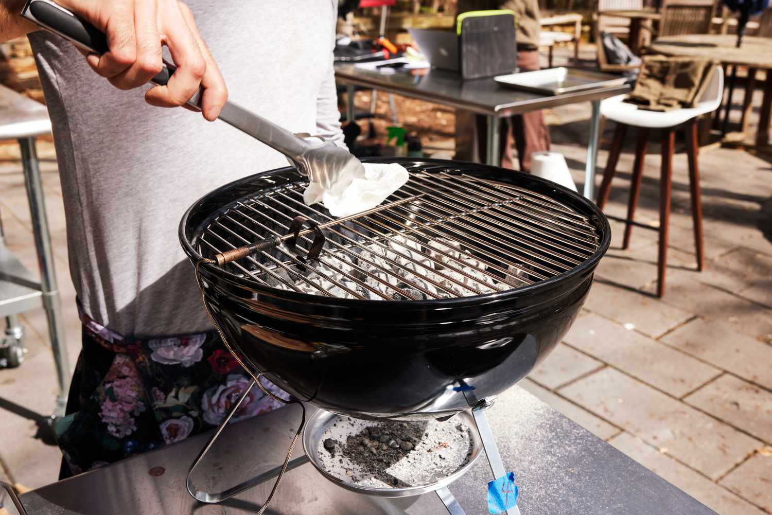 Person cleaning a Weber Smokey Joe Charcoal Grill on a table using tongs with a paper towel 