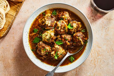 A bowl of French onion slow cooker meatballs topped with melted cheese and chopped parsley, and served with sliced bread and red wine