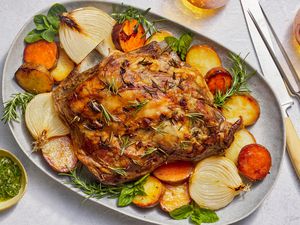 A large platter with a lamb roast served over roasted root vegetables, roasted onion, and a small side bowl of mint sauce