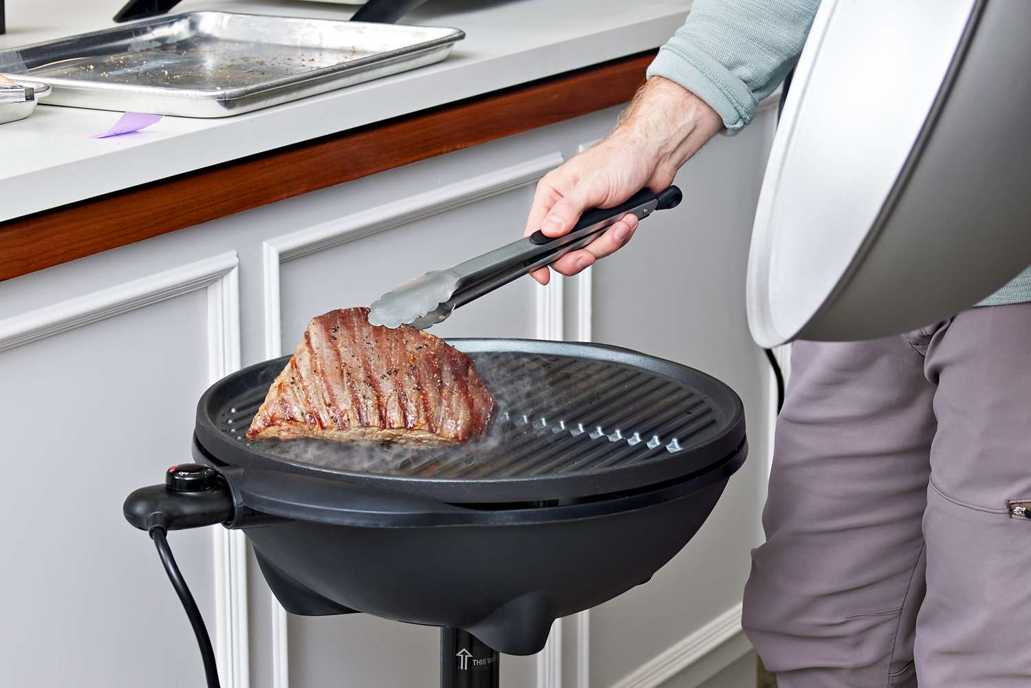 Person holding a flank steak with tongs, next to a George Foreman GFO240S Indoor/Outdoor Electric Grill