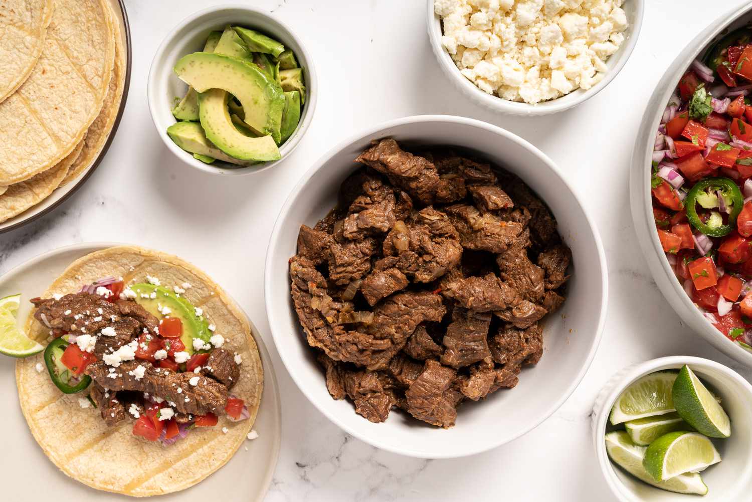 Carne asada in a bowl, with sliced avocado, lime wedges, and other garnishes in bowls 