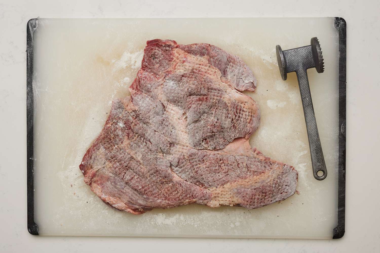 A large, flattened piece of beef lightly coated with flour, on a cutting board with a meat tenderizer 