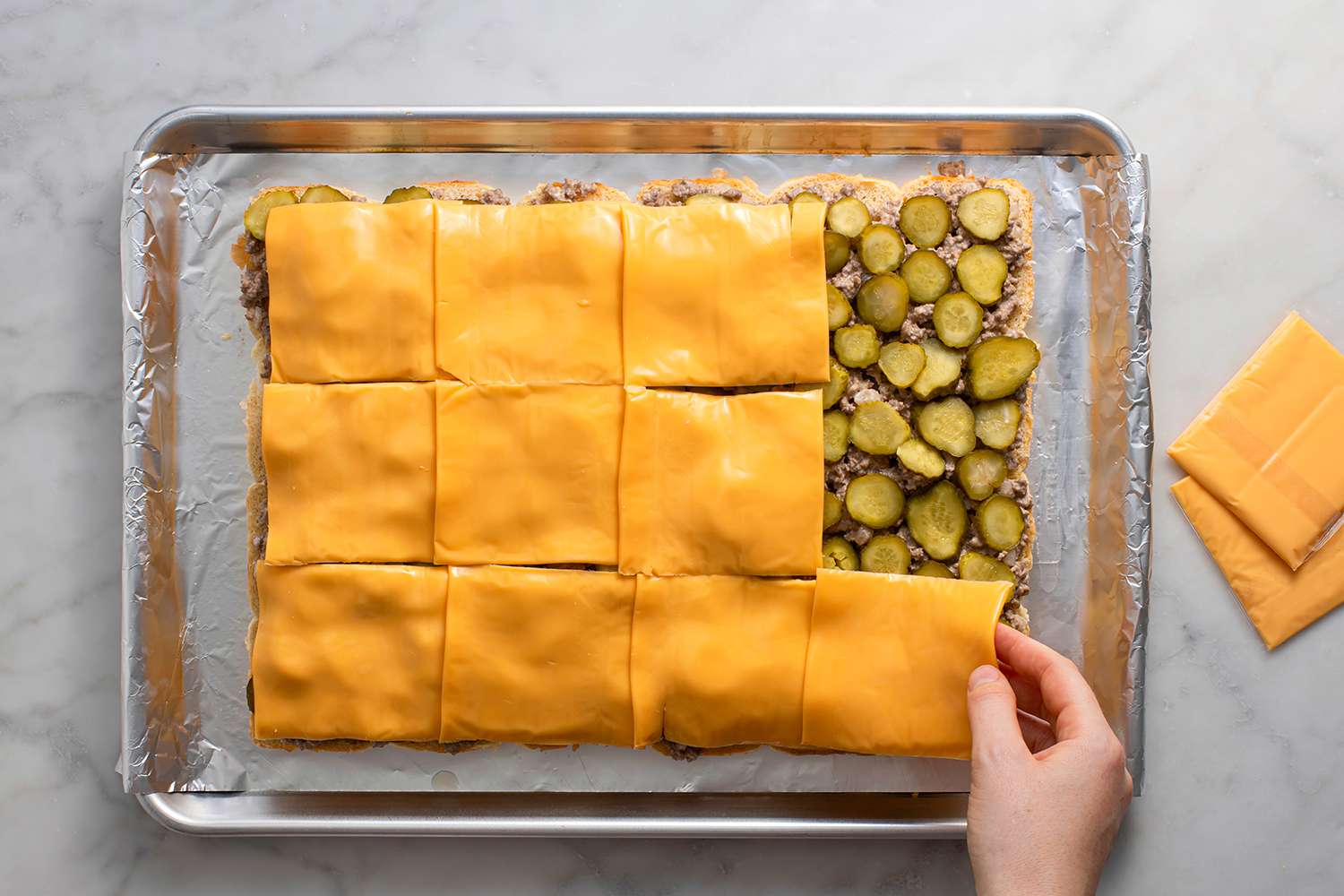 A baking sheet of rolls covered with beef mixture, pickles, and cheese