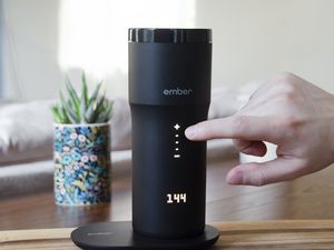 Hand pressing a button on the Ember Travel Mug 2 displayed on a wooden surface next to a potted plant