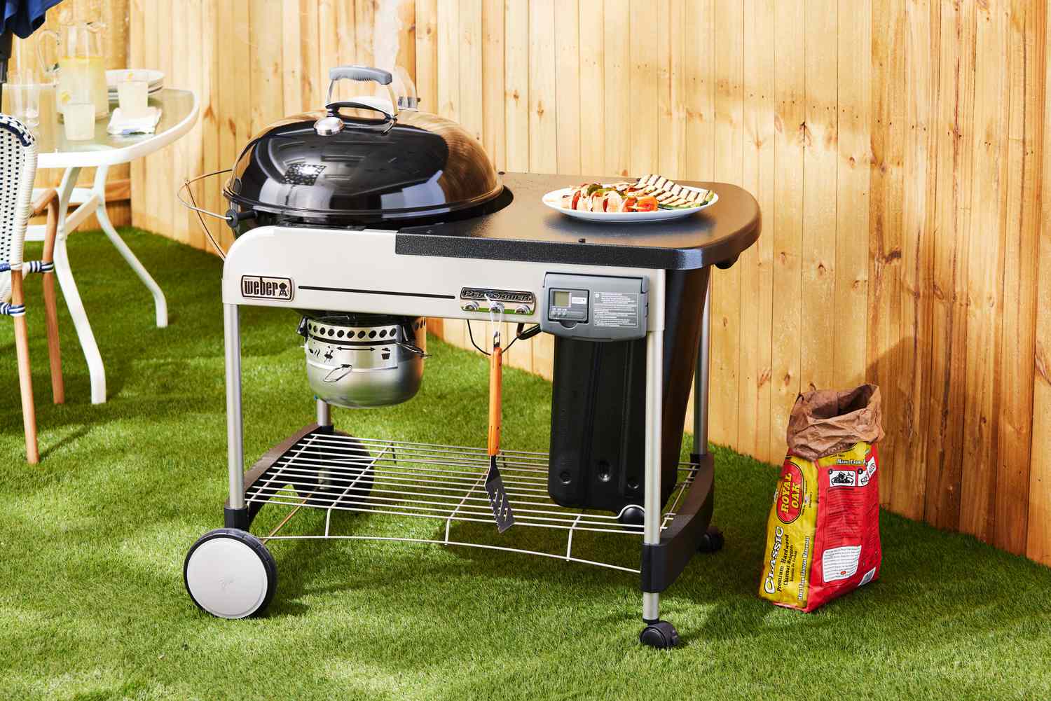 The Weber Performer Deluxe Charcoal Grill on grass near wooden fence