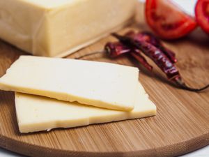 Two slices on white cheese on a bamboo cutting board with a few dried chiles and a quartered Roma tomato in the background