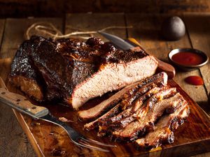 Half a brisket on a wooden cutting board with small pieces cut up in front of it. There is a knife next to it as well as dipping sauce in the background.