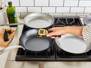 Three carbon steel pans on a gas stove with a hand using a digital thermometer to measure the temperature of one pan 