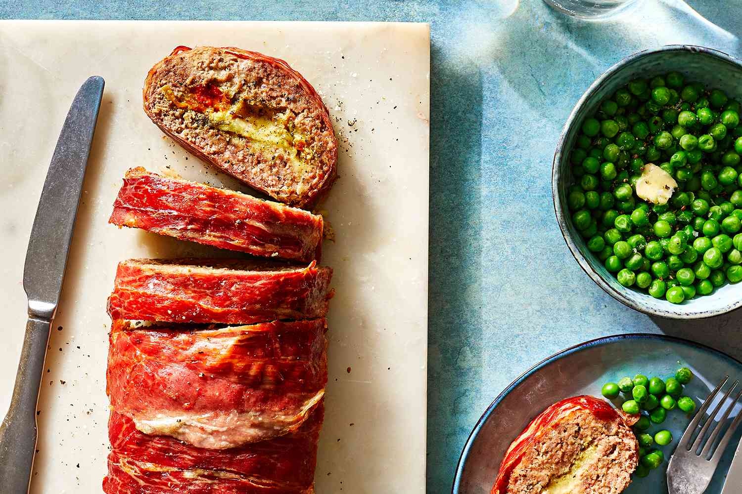 Prosciutto-Wrapped Stuffed Meatloaf on a cutting board with a knife and peas in a bowl 