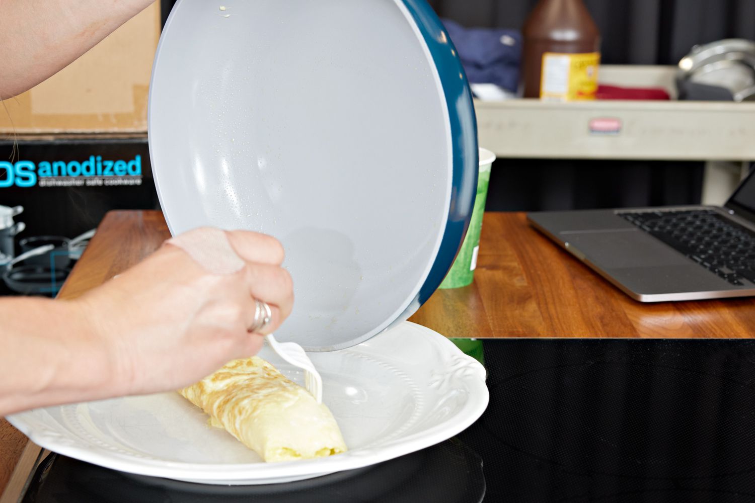 Person's hand sliding omelet onto plate from Caraway Cookware Set frying pan with nearby laptop