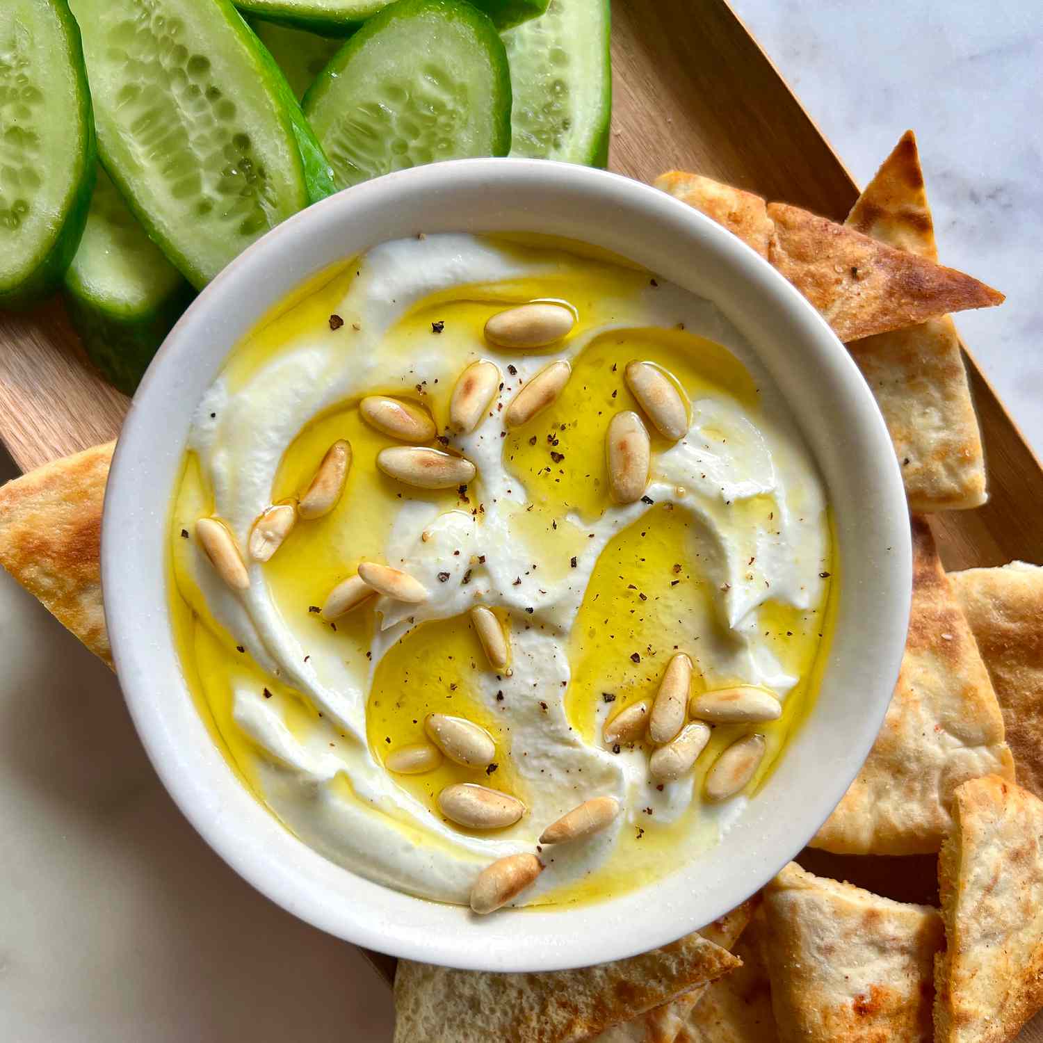 A bowl of creamy whipped feta topped with olive oil and toasted pine nuts on a board with sliced cucumber and pita wedges