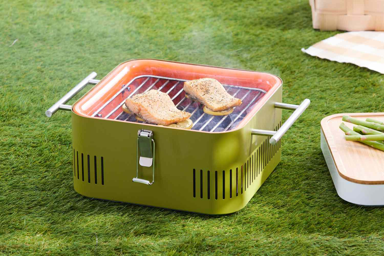 Salmon fillets cooking on lemon slices on a Everdure Cube Portable Charcoal Grill displayed on grass 