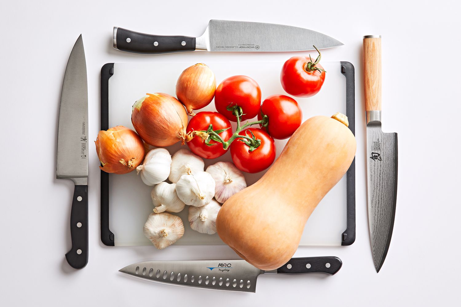 Several chefs knives displayed around a cutting board with vegetables and butternut squash