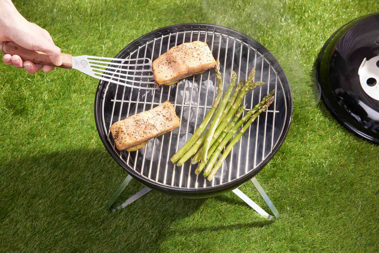 Hand with spatula next to salmon fillets and asparagus on the Weber Smokey Joe Charcoal Grill