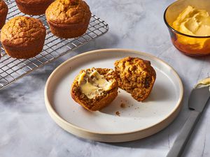 A pumpkin muffin, cut in half with butter, with a cooling rack of pumpkin muffins