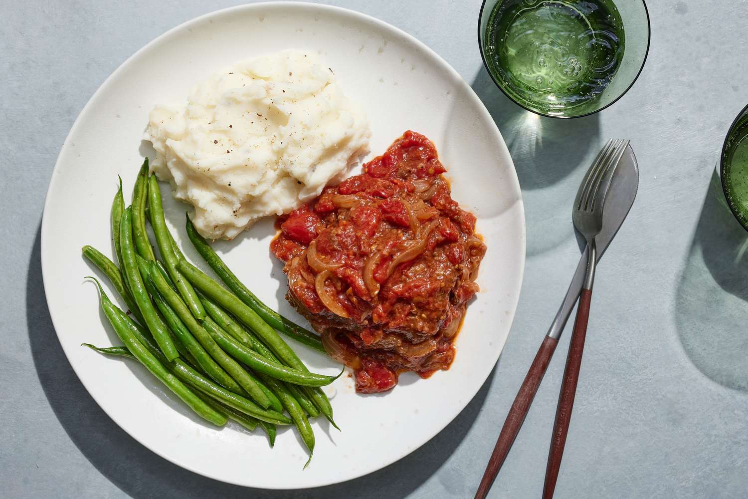 A plate of Swiss steak served with steamed green beans, mashed potatoes, and sparkling water