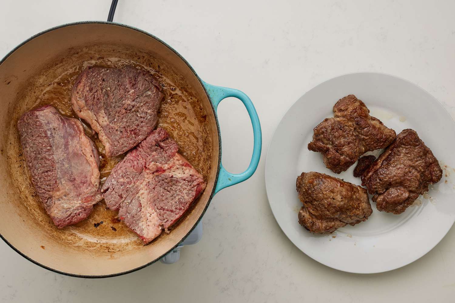 A dutch oven with four three pieces of steak cooking, next to a plate with three pieces of cooked steak