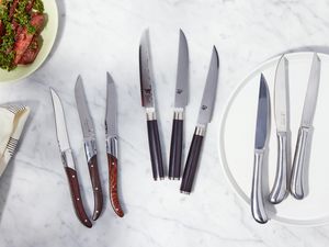 Assortment of the best steak knives we recommend displayed on a white marble countertop next to a plate and a towel