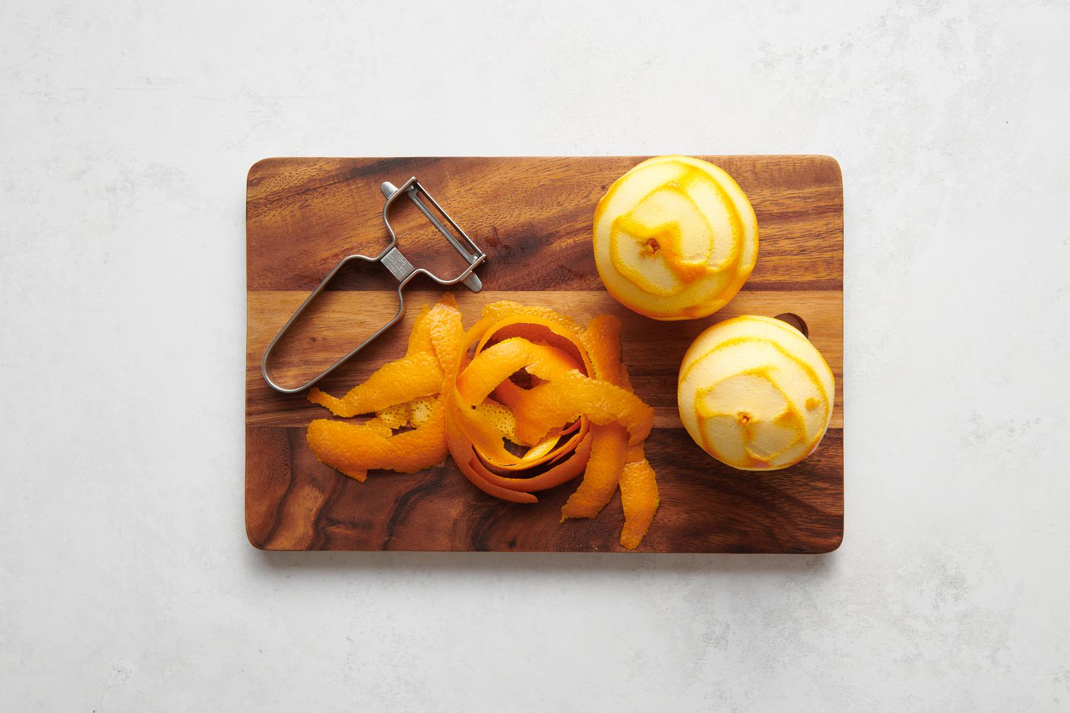 A cutting board with two peeled oranges, a peeler, and a pile of orange peels