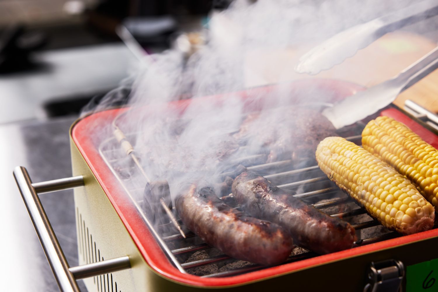 Closeup of sausages, burger and corn cooking on the Everdure Cube Portable Charcoal Grill 