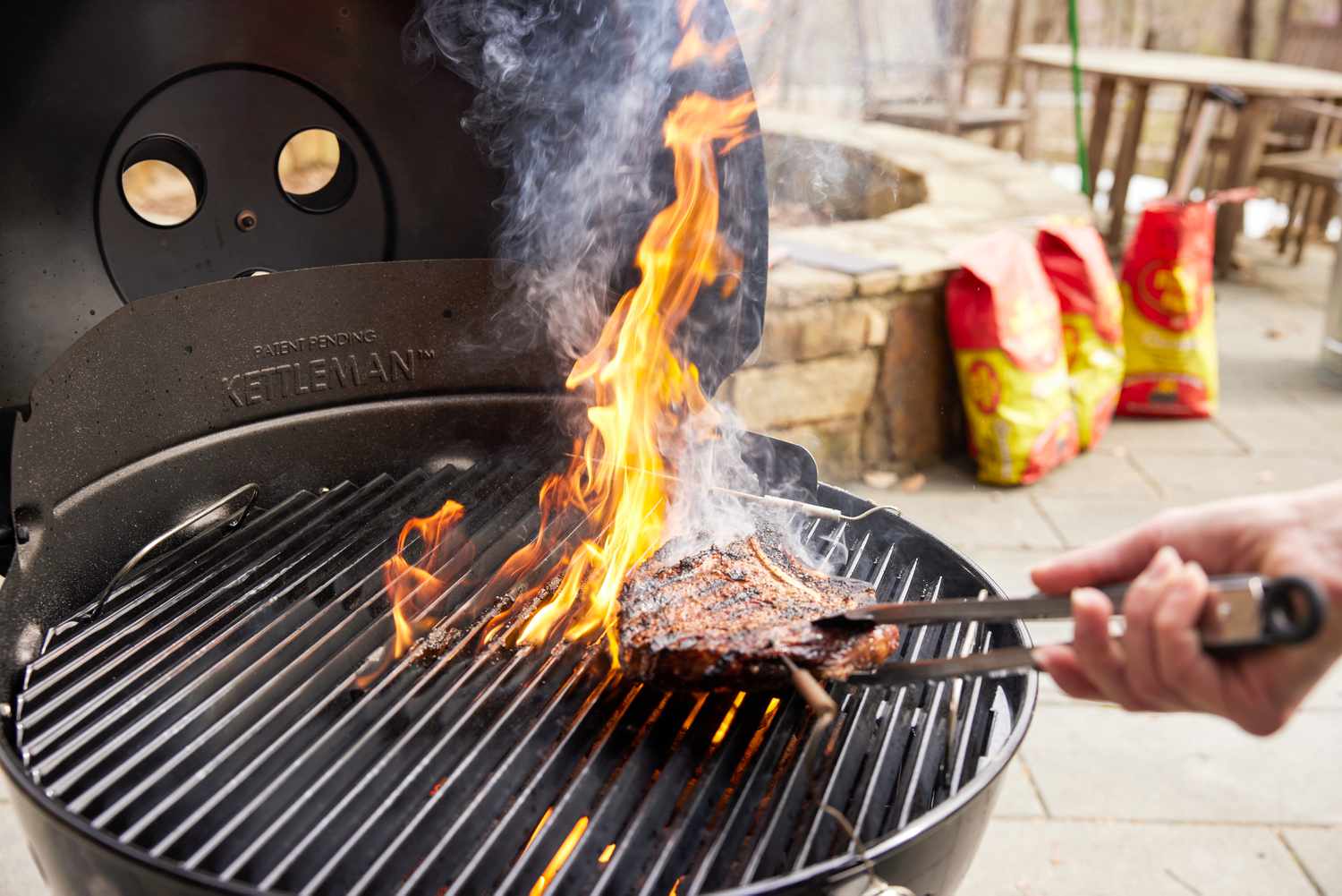 Tongs grasping steak on flaming Char-Broil TRU-Infrared Kettleman Charcoal Grill