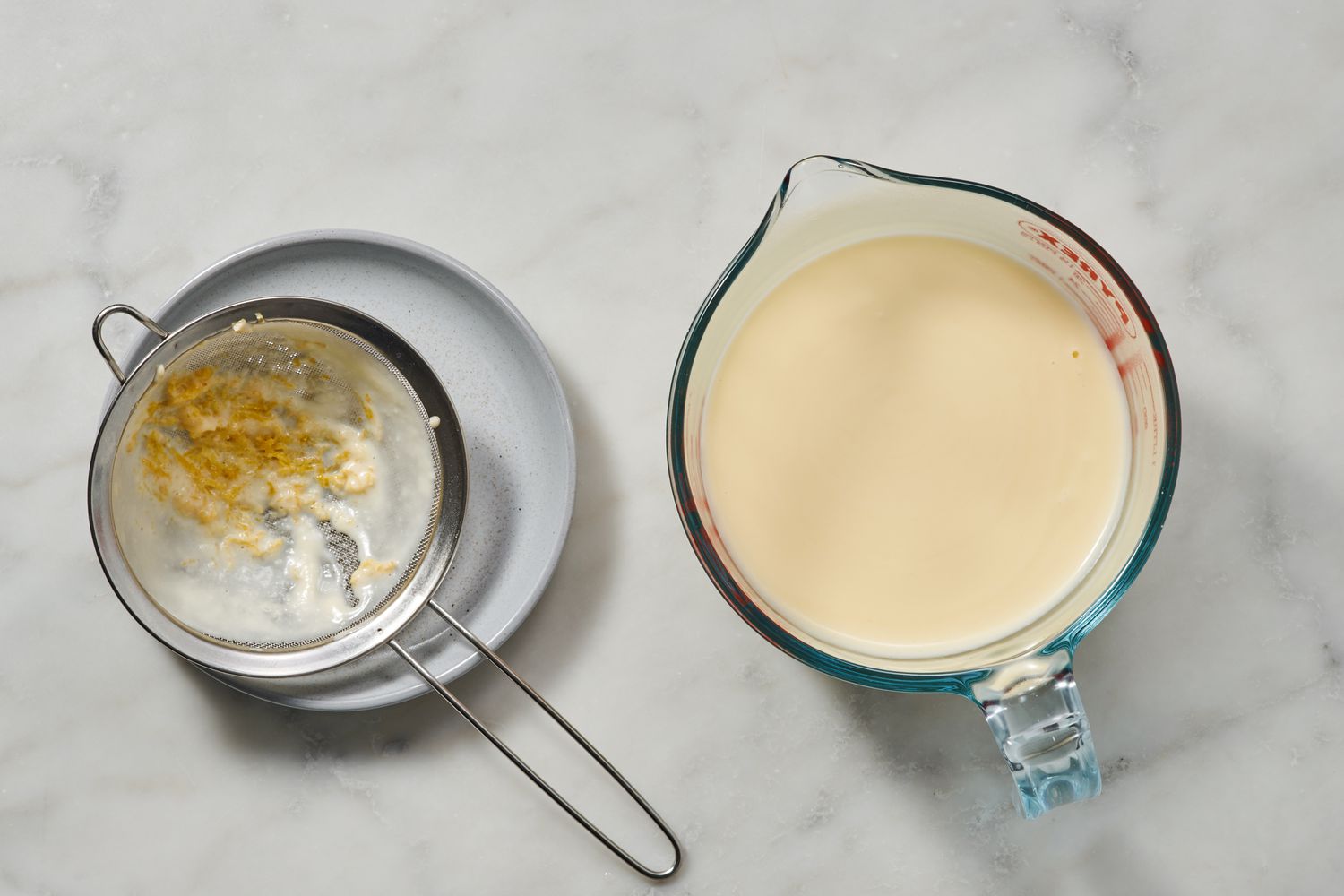 A glass measuring cup of hot cream, lemon juice, and sugar mixture, next to a strainer with lemon zest