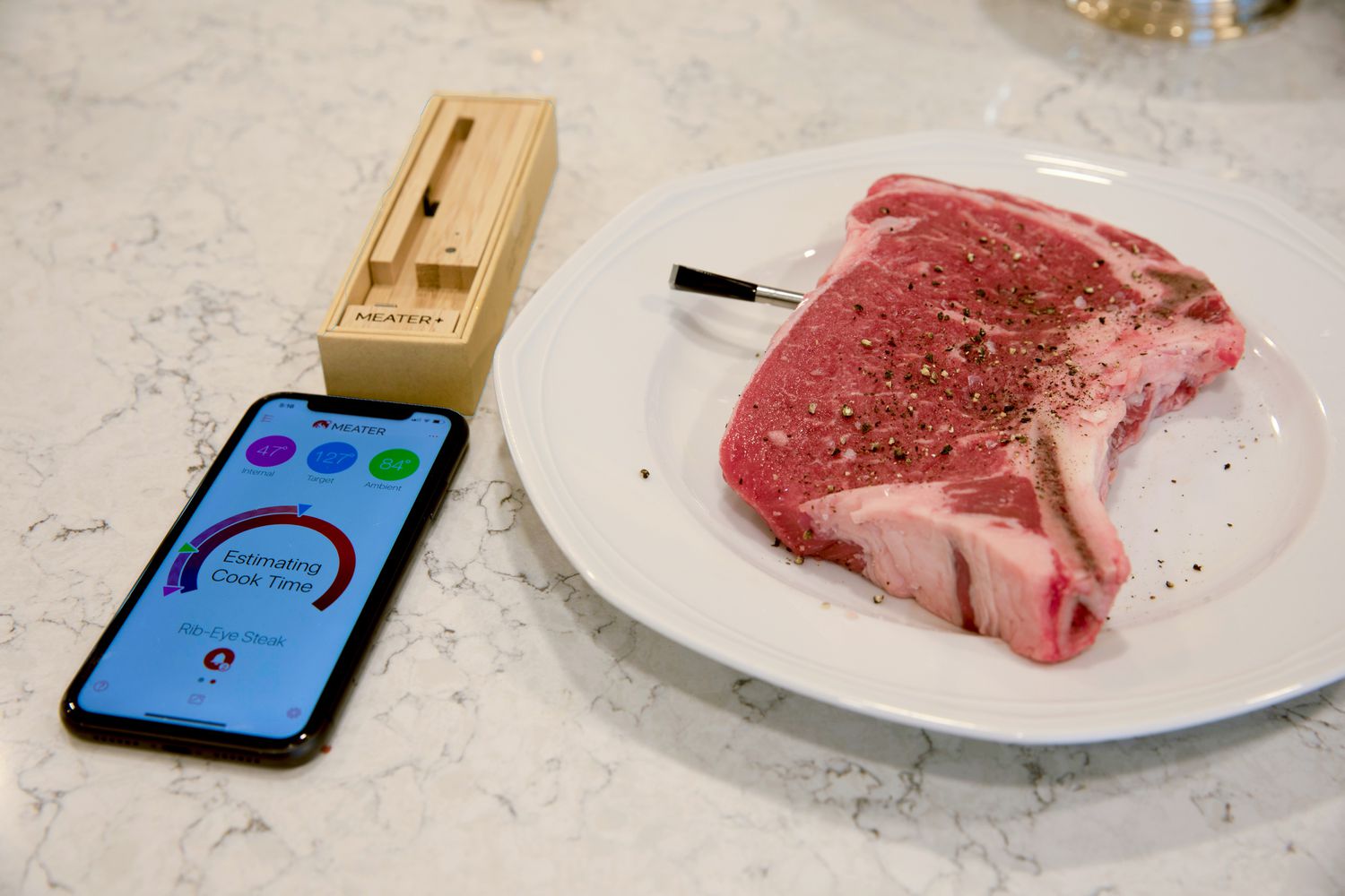 A Meater Plus Thermometer is inserted into a seasoned ribeye. A smart phone shows the estimated cooking time.