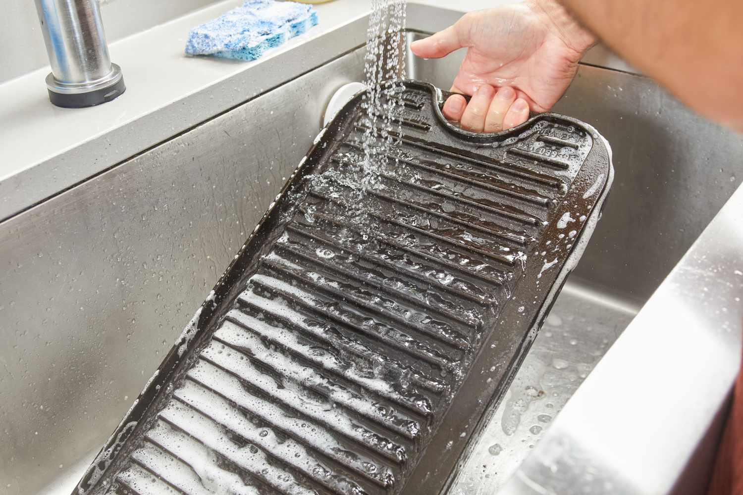 A person washing the Lodge Cast Iron Reversible Grill/Griddle with soap and water.