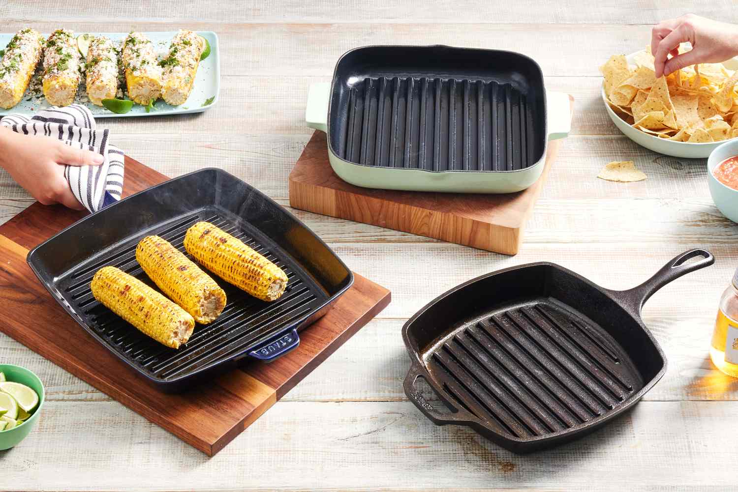 Three grill pans displayed on a wooden table with grilled corn and tortilla trips