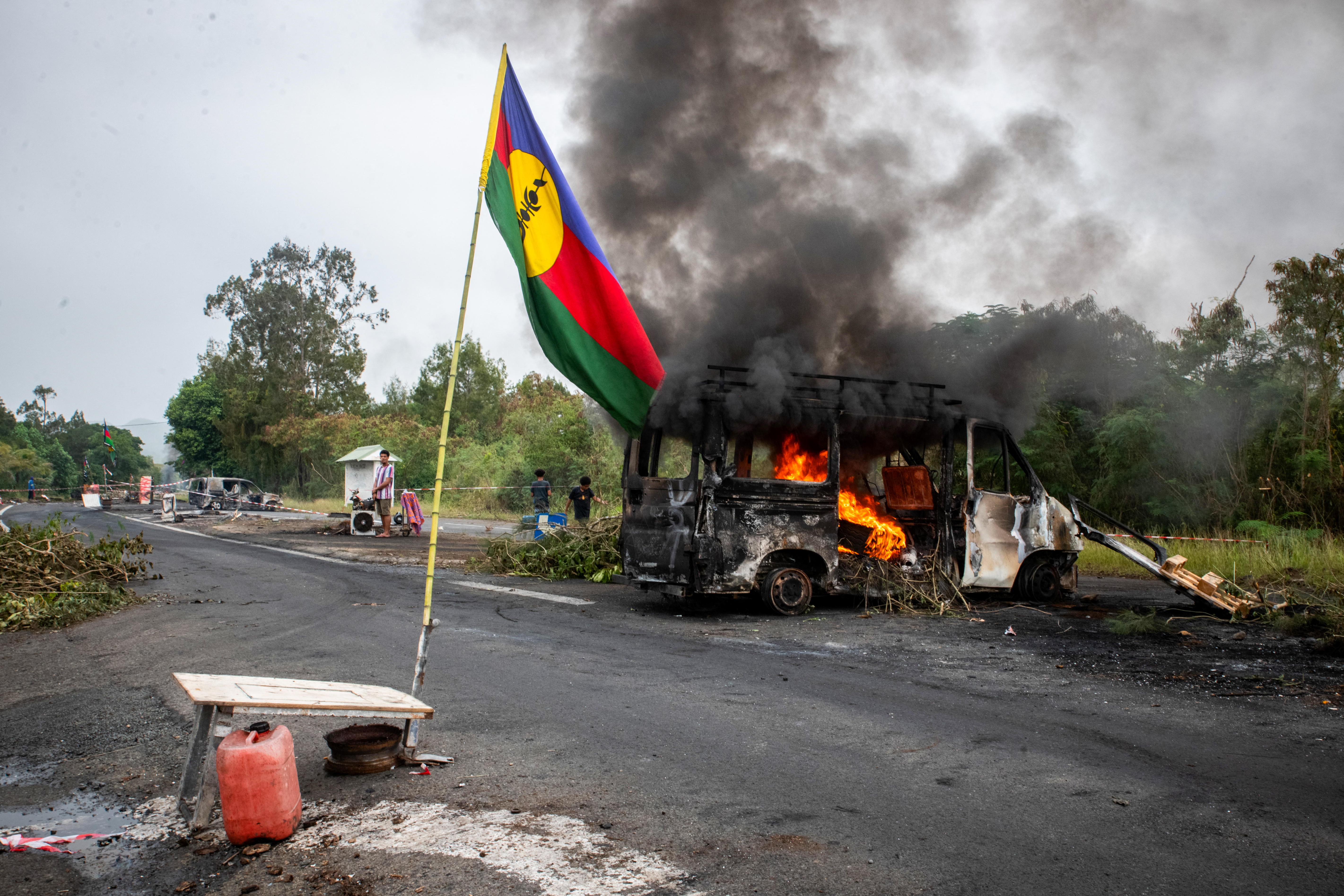 A Kanak flag waves next to a burning vehicle at a roadblock in New Caledonia