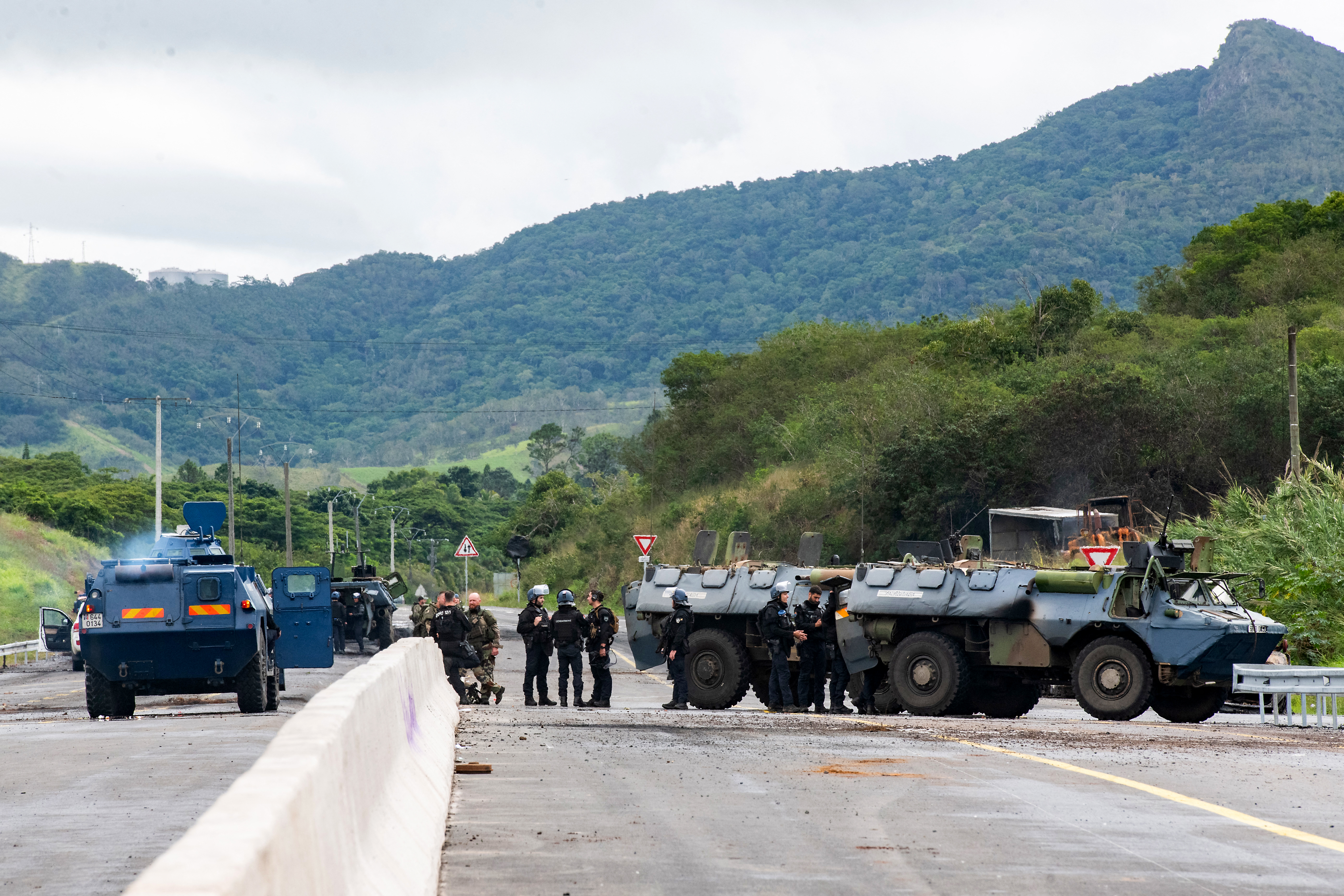 Security officials and armored vehicles of the French Gendarmerie stand guard on the RT1, securing a machine clearing debris and rubbish from a road