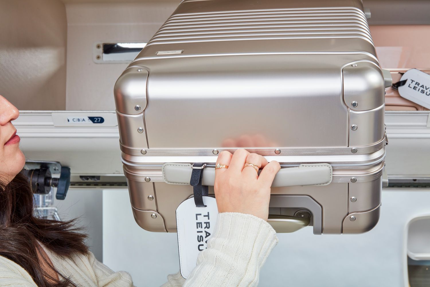 A person places an Arlo Skye The Frame Carry-on Max: Aluminum Edition in an airplane's overhead compartment