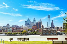 This image shows the skyline of Philadelphia, demonstrating the variety in the structures ranging from century-old buildings to modern structures. The Delaware River bisects the image in the lower third, and a small park fills the immediate foreground.