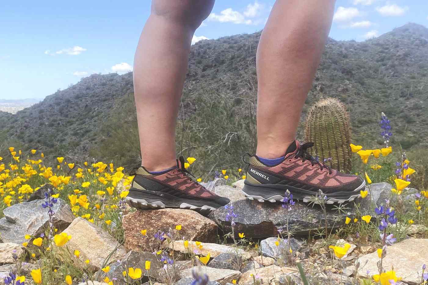 A side profile of a woman wearing Merrell Speed Eco Hiking Shoes while standing on rocks among yellow and purple flowers