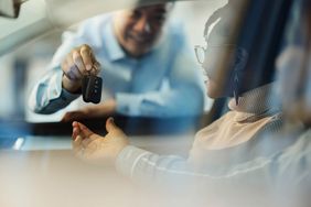 Close up of a salesman giving new car keys to his black customers in a car showroom. The view is through glass.