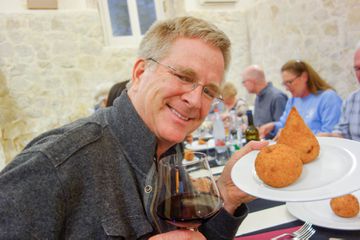 Rick Steves smiling with a plate of fried snacks and a glass of red wine in Italy