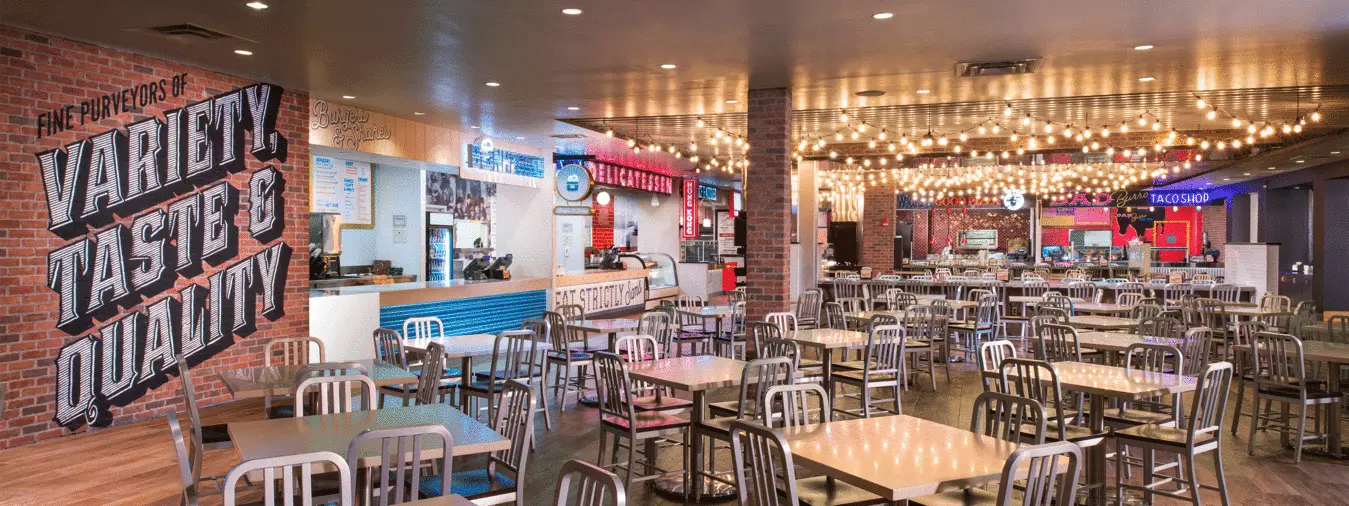 Multiple empty tables and chairs with bright lights above, near food stands