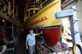 Tom Sweeney stands next to a large yellow and red wooden boat that's up on blocks in a workshop.