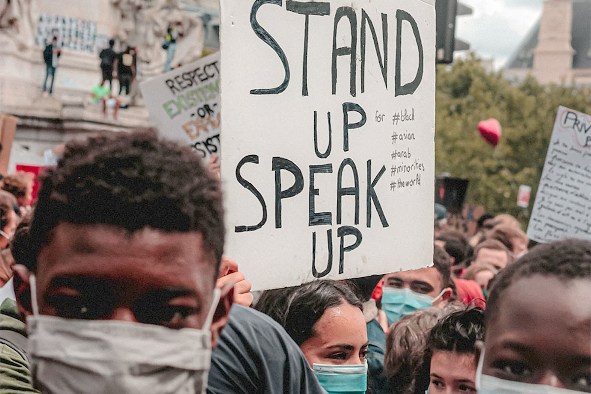 Protestors hold up a sign that reads: Stand Up Speak Up. 